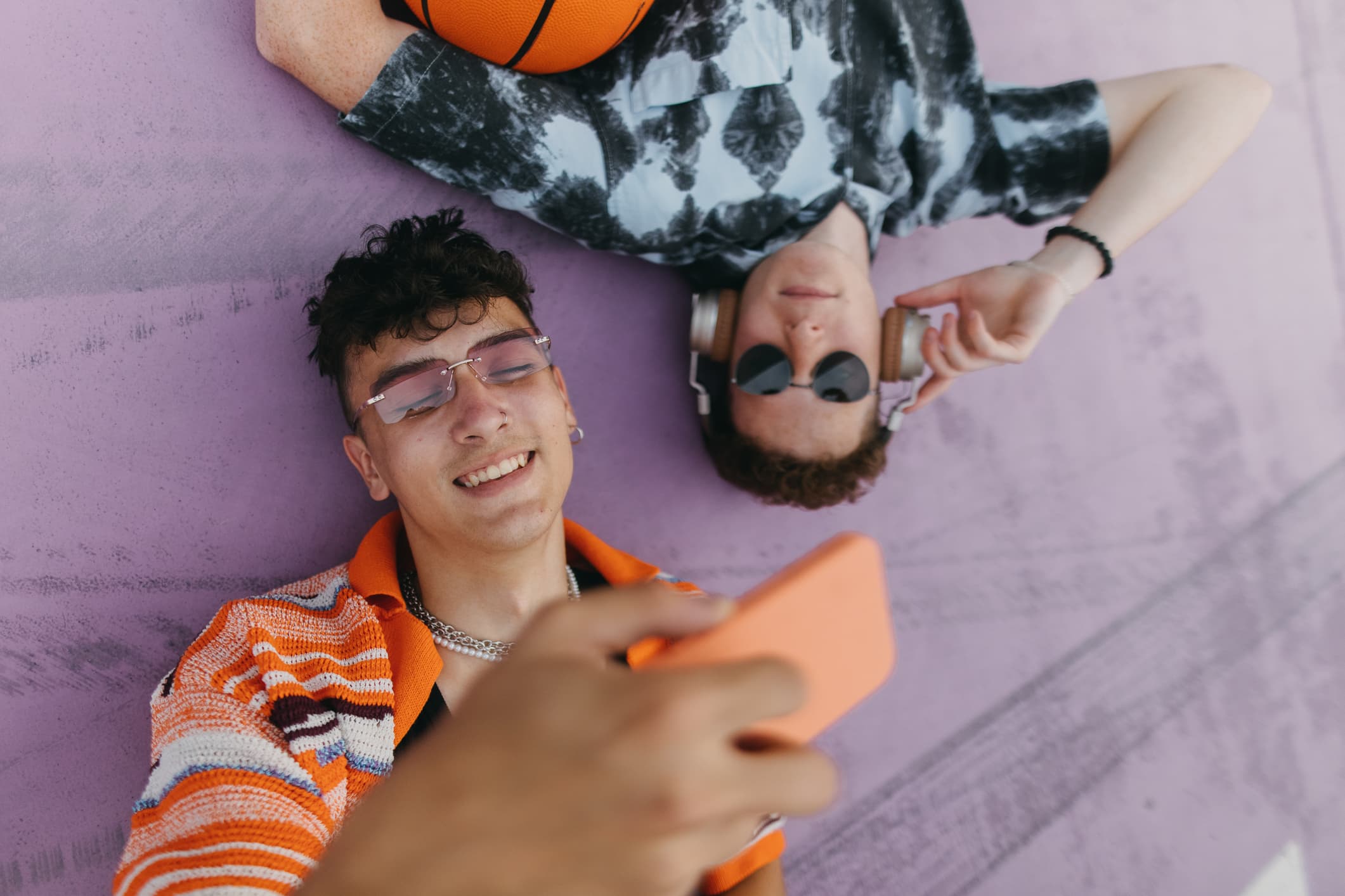 Portrait of young stylish boy zoomers lying on the concrete floor watching social media content on smartphone. Generation z friends hanging out together outdoors in the city, spending time online. Concept of power of friendship and social strength of gen Z.
